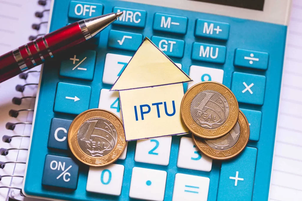 Imagem de uma calculadora azul com moedas, uma caneta vermelha e um recorte de papel no formato de uma casa com a inscrição "IPTU" colocados sobre as teclas