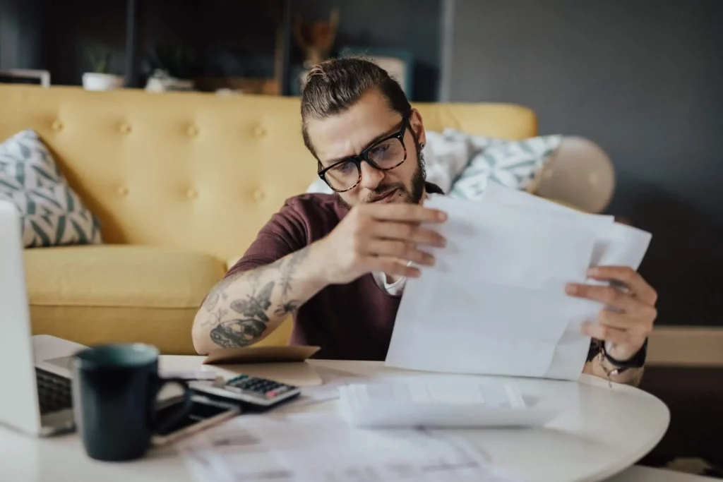 Imagem de um homem de óculos e tatuagens sentado no chão em frente a um sofá amarelo. Um laptop, uma calculadora, uma caneca e mais papéis estão sobre a mesa à sua frente