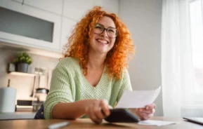 Imagem de uma mulher sorridente, de cabelos ruivos encaracolados e óculos, sentada na mesa de uma cozinha bem iluminada, com a luz do sol entrando pela janela, com uma calculadora e um papel na mão