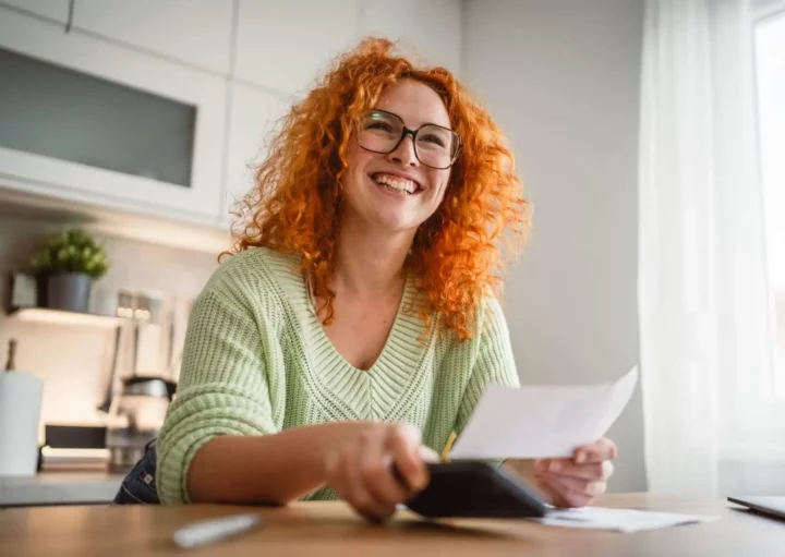 Imagem de uma mulher sorridente, de cabelos ruivos encaracolados e óculos, sentada na mesa de uma cozinha bem iluminada, com a luz do sol entrando pela janela, com uma calculadora e um papel na mão