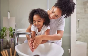 Imagem de uma mãe ajudando a filha a lavar as mãos em uma pia de banheiro para ilustrar matéria sobre a troca de titularidade Sabesp