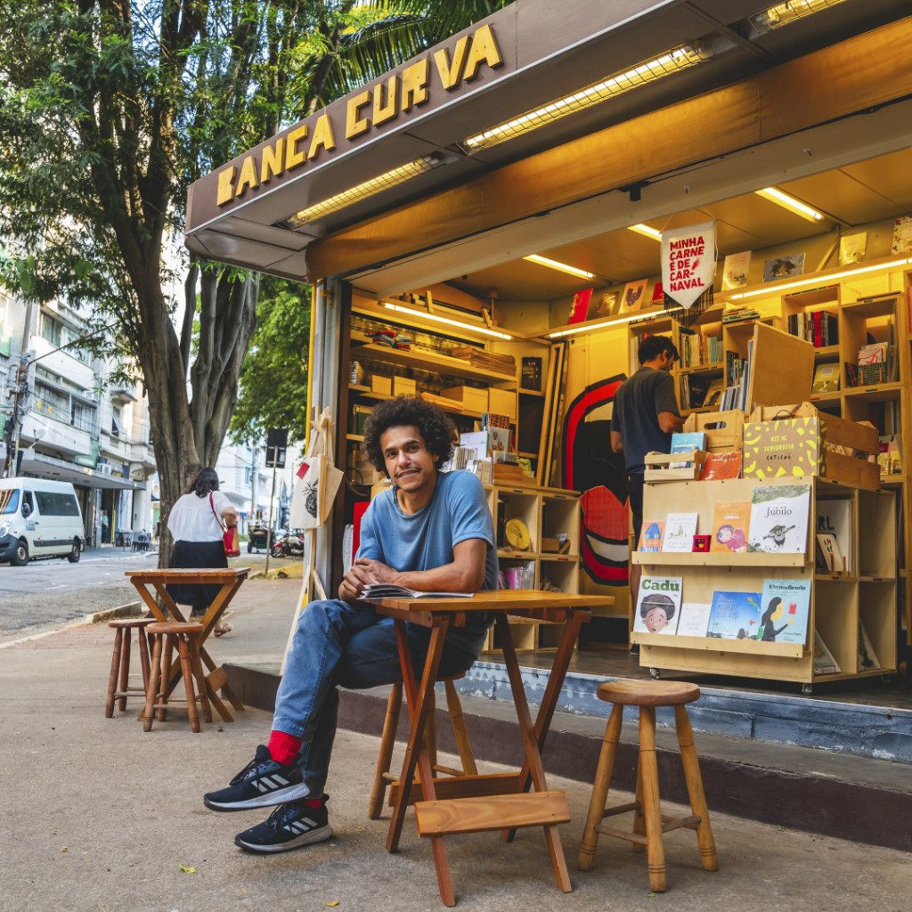 Foto do Tom Vieira sentado em uma mesa de madeira na Banca Curva