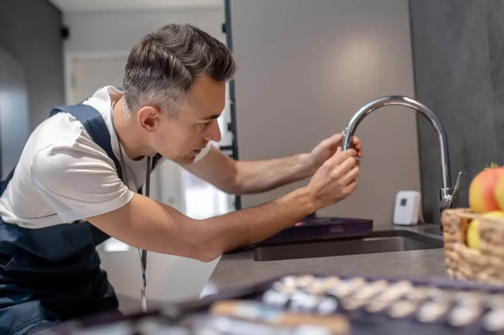 Imagem de um homem de macacão de trabalho fazendo a vistoria de uma torneira de cozinha usando ferramentas e concentrando-se atentamente na tarefa. Uma caixa de ferramentas e uma cesta com frutas estão visíveis no balcão próximo.