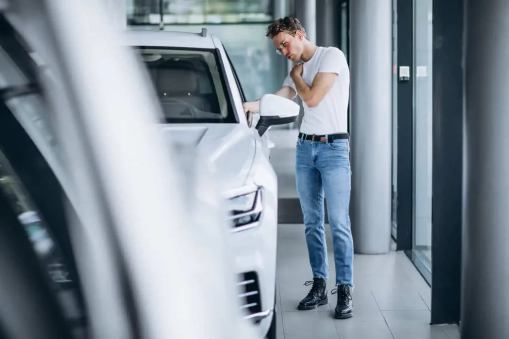 Imagem de um homem de calça jeans e camiseta branca está ao lado de um carro branco examinando as características do veículo e da vaga para ilustrar matéria sobre vistoria de imóvel locado.
