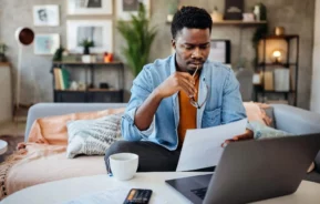 Imagem de um homem sentado no sofá de casa em frente a uma mesa de centro onde estão uma xícara de café e um notebook para ilustrar matéria sobre a pergunta: financiar um imóvel vale a pena?