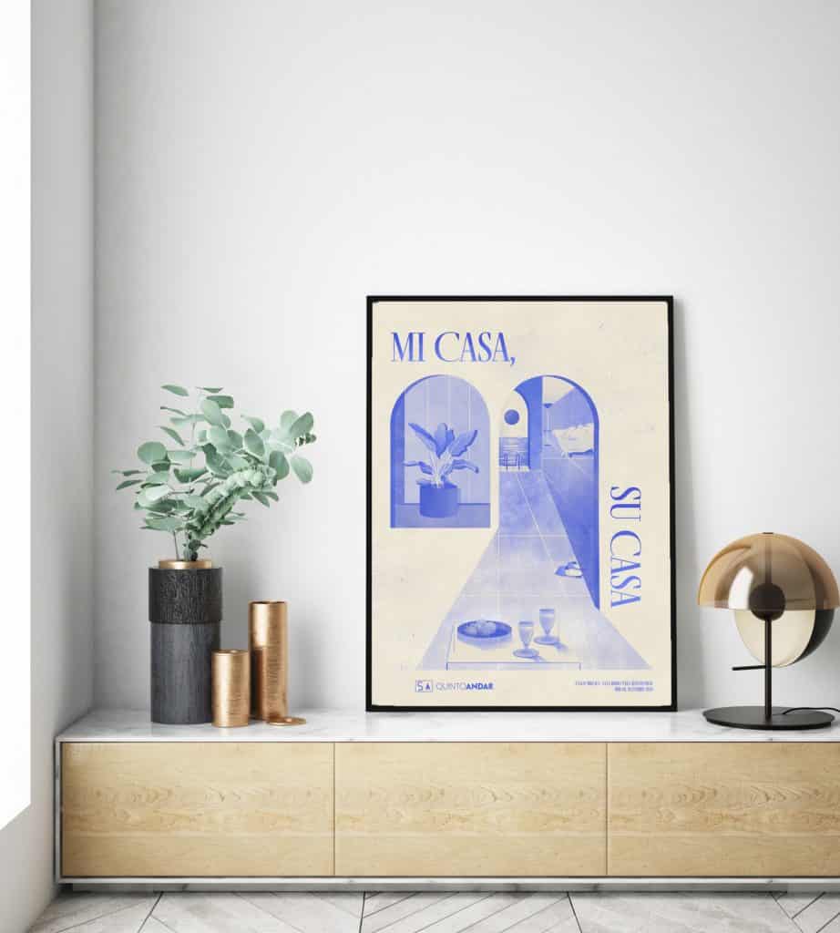 Um interior moderno com um armário de madeira clara, planta em vaso, vasos metálicos e uma luminária de mesa dourada. Um pôster emoldurado na parede diz "Mi Casa, Su Casa" em azul, com uma cena ilustrada — estilizado por QuintoAndar Colab.