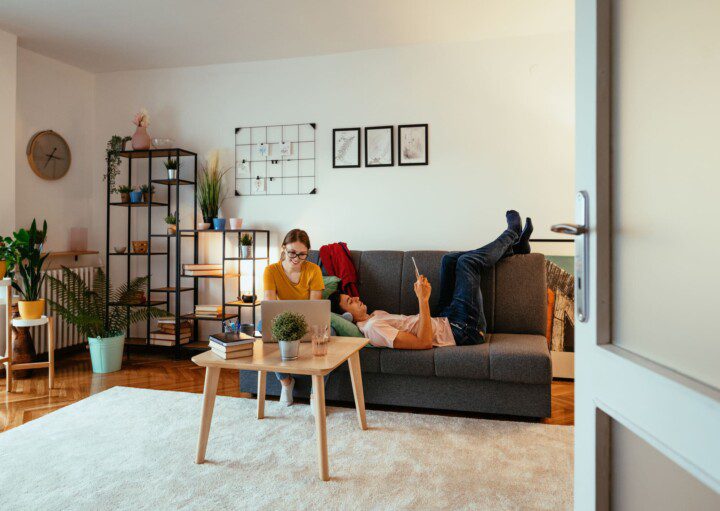 Foto que ilustra matéria sobre apartamento mobiliado para alugar mostra uma mulher e um homem em uma sala com quadros e plantas. A mulher mexe em um notebook, enquanto o homem está deitado no sofá.