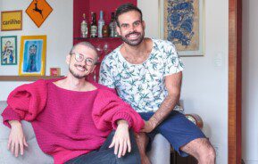 Casal formado por dois homens, sentados em um sofá e sorrindo, em um apartamento no Edifício Saint Honoré, na Avenida Paulista, em São Paulo