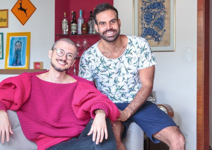 Casal formado por dois homens, sentados em um sofá e sorrindo, em um apartamento no Edifício Saint Honoré, na Avenida Paulista, em São Paulo