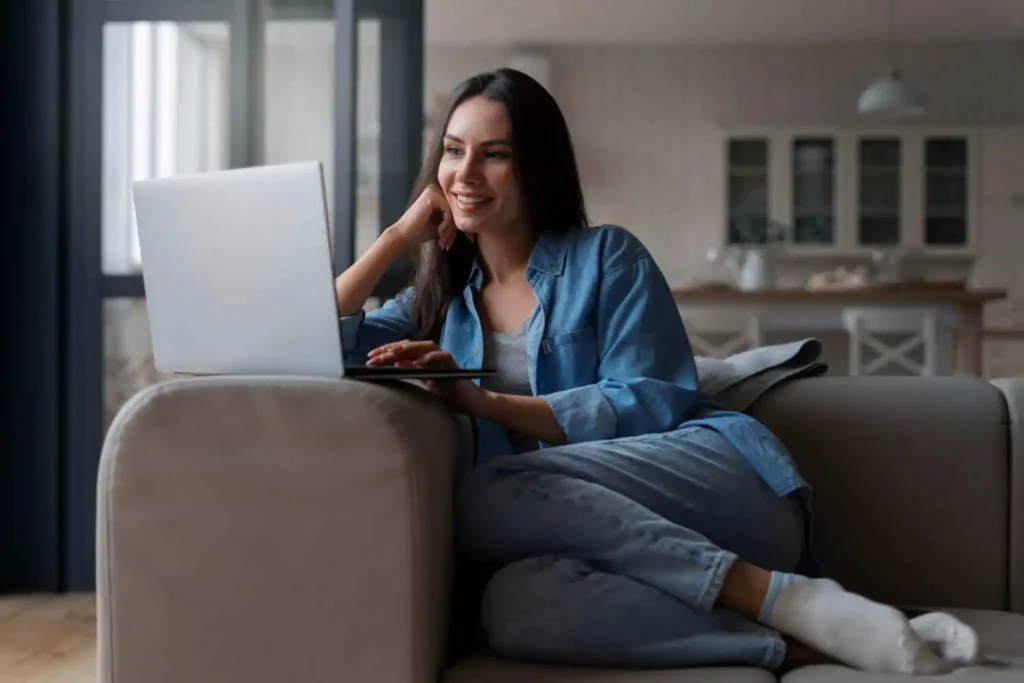 Imagem de uma mulher de camisa jeans e calça jeans sentada confortavelmente em um sofá, sorrindo enquanto usa seu laptop para navegar em uma imobiliária digital em uma sala de estar moderna e aconchegante.