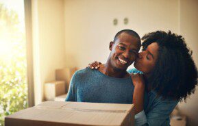 Foto que ilustra matéria sobre proposta de locação mostra um casal jovem que acaba de se mudar para uma casa nova. Na imagem, o homem sorridente ganha um beijo de sua parceira na bochecha.