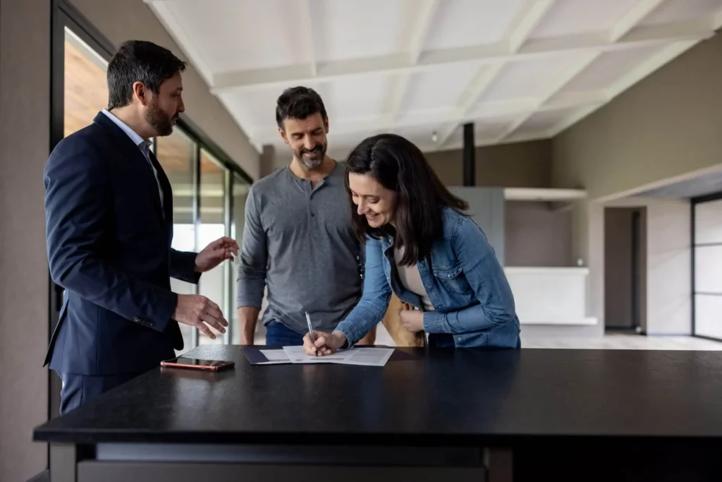 Imagem de uma mulher assinando um documento que está em cima da bancada de um imóvel vazio ao lado do marido e de um corretor de imóveis para ilustrar matéria sobre quem paga a corretagem de imóveis