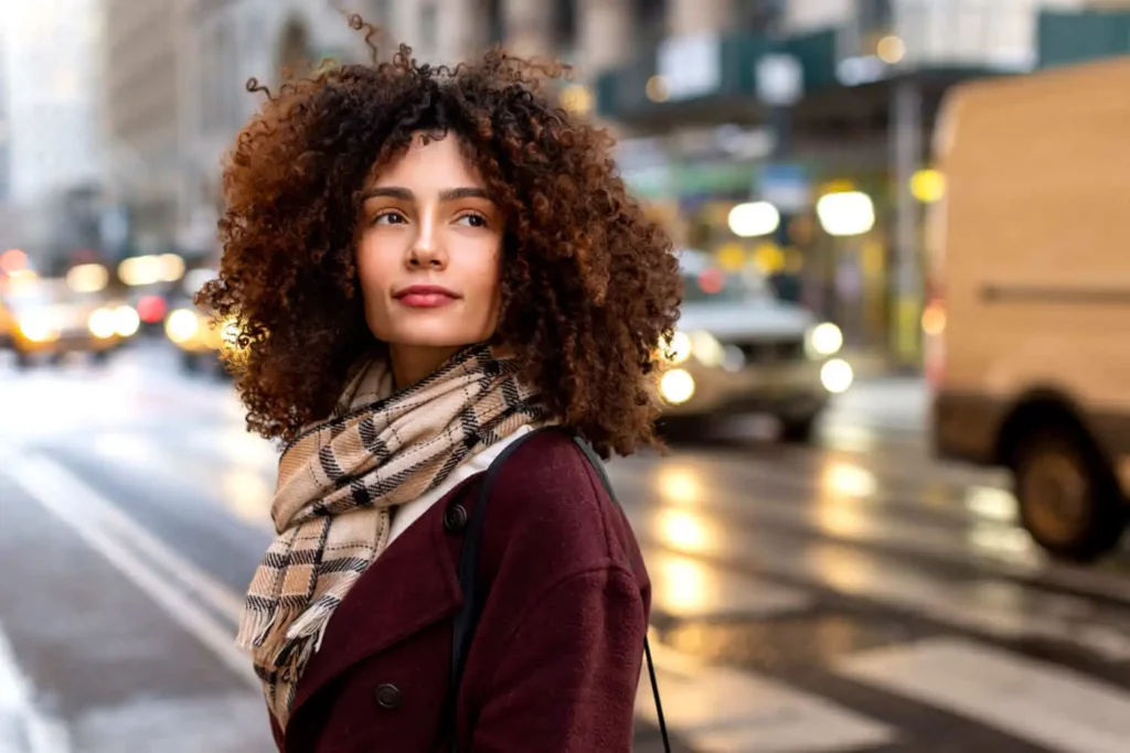  Imagem de uma mulher de cabelo cacheado usando casaco e cachecol atravessando a faixa de pedestre enquanto carros passam pela rua para ilustrar matéria sobre os bairros nobres de São Paulo