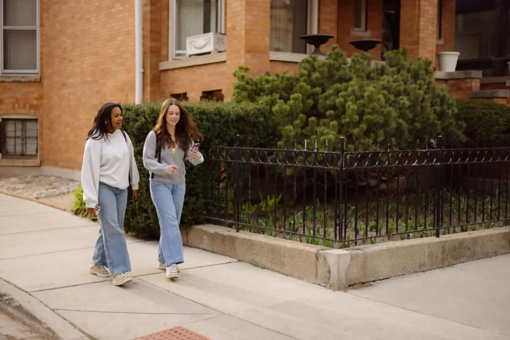Imagem de duas mulheres jovens caminhando e conversando em uma calçada ao lado de um prédio de tijolos e arbustos verdes. Ambas vestem roupas casuais com jeans e tênis, e uma delas está segurando um telefone.