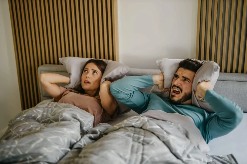 Imagem de um casal deitado na cama segurando um travesseiro nas orelhas para ilustrar matéria sobre como funciona a Lei do Silêncio