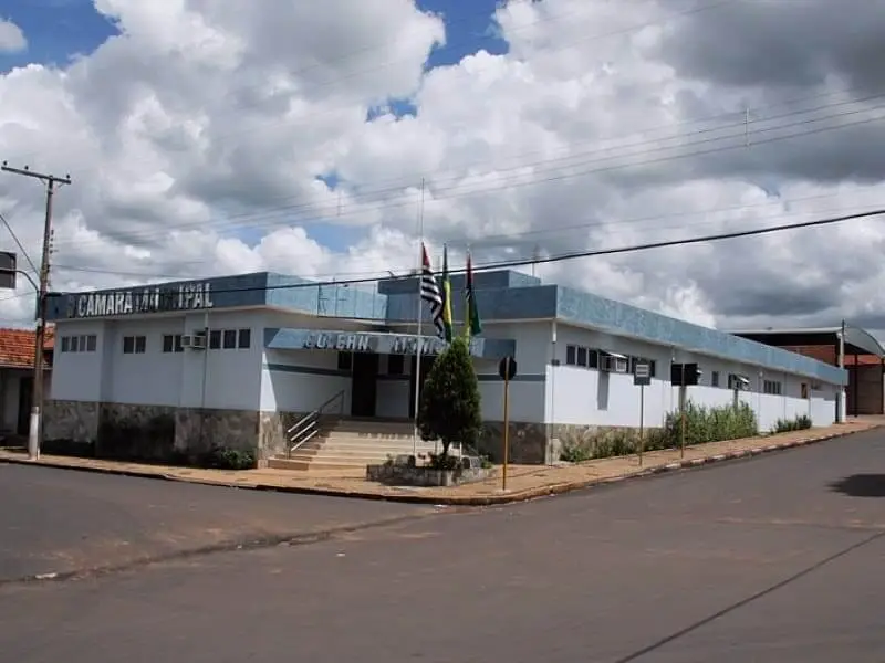 Imagem da Câmara Municipal de Cândido Rodrigues, em São Paulo, mostra prédio branco de um andar com detalhes em azul em uma esquina sob um céu nublado. Três mastros de bandeira estão na entrada, com uma pequena árvore ao lado da escada