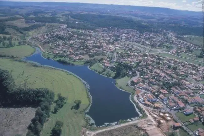 Imagem da vista aérea do Lago das Palmeiras, em Águas de São Pedro, em São Paulo, mostra o lago curvando-se através de campos verdes e casas densamente povoadas, cercadas por árvores e colinas onduladas sob um céu parcialmente nublado