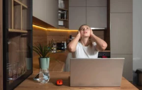Imagem de uma mulher em casa sentada na mesa com um notebook logo em frente, tapando os ouvidos com as mãos e olhando para cima para ilustrar matéria sobre o que é a Lei do Silêncio