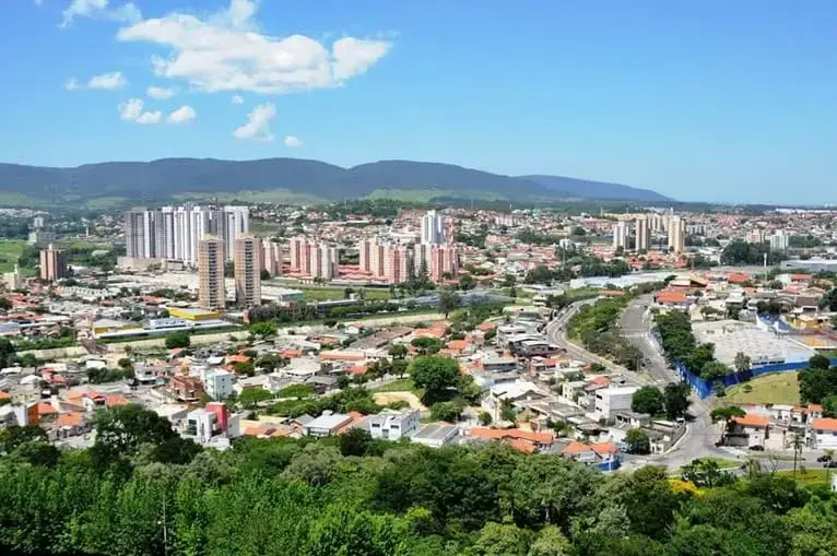 Imagem da vista panorâmica de uma cidade de Jundiaí, em São Paulo, com aglomerados de apartamentos altos, casas menores, estradas e vegetação em um cenário de montanhas e um céu azul com algumas nuvens