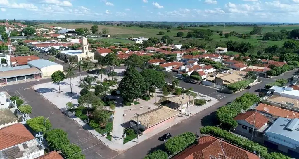 Imagem da vista aérea da cidade de Gabriel Monteiro, em São Paulo, mostra uma praça central, telhados vermelhos, uma torre de igreja, árvores exuberantes e campos verdes sob um céu azul com nuvens dispersas