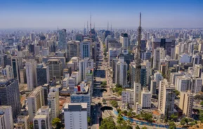 Imagem da vista aérea da Avenida Paulista, em São Paulo, mostra vários prédios altos entrecortados por ruas e avenidas movimentadas, sob um céu azul claro.