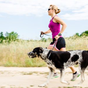 Uma mulher com roupas esportivas e &oacute;culos de sol corre ao ar livre em uma trilha de terra, segurando uma guia presa a um grande cachorro preto e branco com focinheira. Grama e &aacute;rvores s&atilde;o vis&iacute;veis ao fundo.