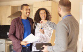 Em foto que ilustra matéria sobre aluguel sem fiador, um casal sorridente aparece. O homem segura papéis enquanto a mulher recebe de um homem de terno posicionado de costas as chaves de um imóvel