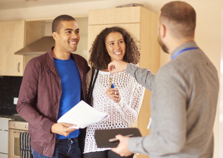 Em foto que ilustra matéria sobre aluguel sem fiador, um casal sorridente aparece. O homem segura papéis enquanto a mulher recebe de um homem de terno posicionado de costas as chaves de um imóvel