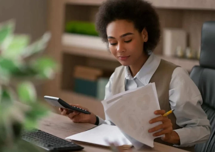 Imagem de uma mulher negra sentada em uma mesa, segurando uma calculadora e analisando documentos sobre alugar ou financiar, com um caderno aberto e um lápis à sua frente, parecendo concentrada.