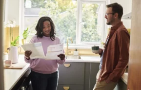 Imagem de um casal na cozinha de uma casa enquanto a mulher olha para algumas contas para ilustrar matéria sobre como declarar imóvel financiado no Imposto de Renda