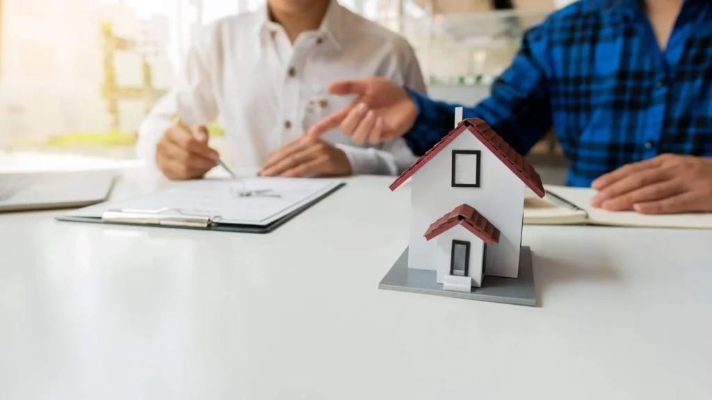 Imagem de duas pessoas sentadas em uma mesa onde tem uma prancheta com folhas e uma miniatura de uma casa em cima para ilustrar matéria sobre como declarar imóvel financiado nunca declarado