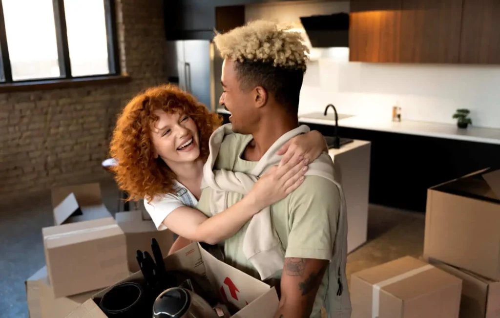 Imagem de um casal sorridente se abraça enquanto desempacota caixas em uma cozinha moderna, sugerindo que eles estão se mudando para uma nova casa juntos, possivelmente um imóvel de leilão.