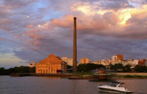 Foto que ilustra matéria sobre os melhores bairros de Porto Alegre mostra a Usina do Gasômetro, um dos pontos turísticos da cidade, às margens do Rio Guaíba, ao entardecer. Além do prédio e da chaminé ao lado, uma lança passa à frente.