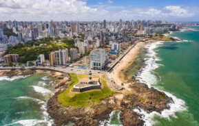 Foto que ilustra matéria sobre os melhores bairros de Salvador mostra os prédios do bairro da Barra vistos do alto, com o Farol da Barra e pedaços da praia em primeiro plano.