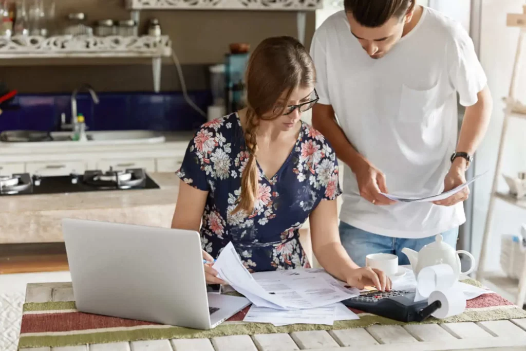 Imagem de uma mulher com um vestido floral e óculos olhando para papéis e usando uma calculadora, enquanto conversa com um homem de camisa branca segurando documentos. Eles trabalham juntos em uma mesa de cozinha com um laptop e um bule de chá.