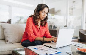 Em foto que ilustra o quiz “Você sabe quanto custa essa casa?”, uma mulher sentada em um sofá usa um laptop posicionado em uma mesa de centro, ao lado de um bloco de anotações com uma caneta em cima.