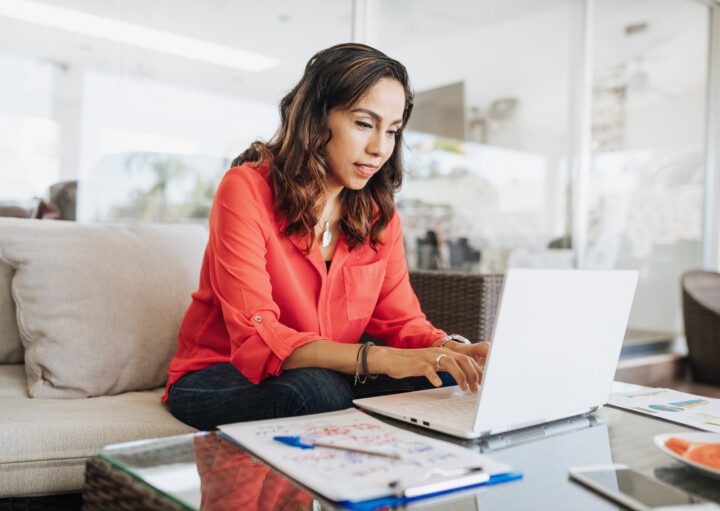 Em foto que ilustra o quiz “Você sabe quanto custa essa casa?”, uma mulher sentada em um sofá usa um laptop posicionado em uma mesa de centro, ao lado de um bloco de anotações com uma caneta em cima.