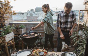 A foto mostra uma área gourmet pequena. Há na imagem: um homem e uma mulher fazendo churrasco. Tem também uma churrasqueira, uma mesa com comidas e plantas decorativas.