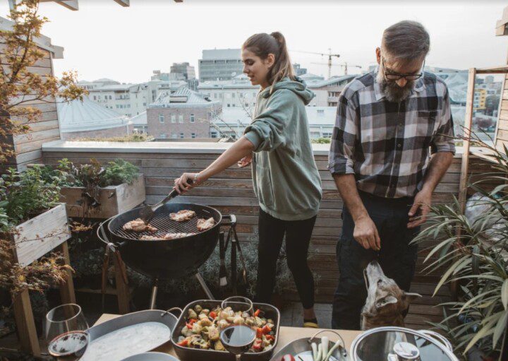 A foto mostra uma área gourmet pequena. Há na imagem: um homem e uma mulher fazendo churrasco. Tem também uma churrasqueira, uma mesa com comidas e plantas decorativas.