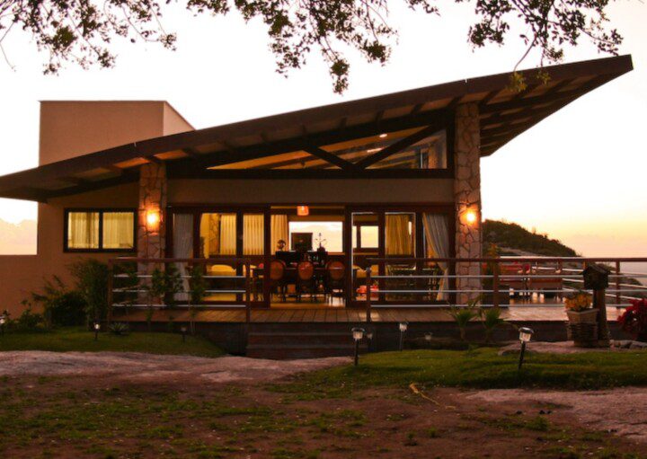 Foto que ilustra matéria sobre casas rústicas mostra uma casa no alto de uma estrutura rochosa, ao entardecer. A construção é cercada por uma varanda, tem um telhado em diagonal e uma bela vista para o pôr do sol.