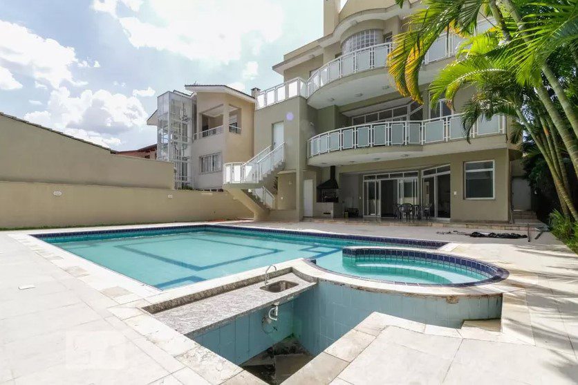 Casa de Luxo em Condomínio em Santana de Parnaíba, com uma grande piscina e a casa aos fundos, com três andares. Há, ainda, uma palmeira à direita encobrindo levemente a imagem