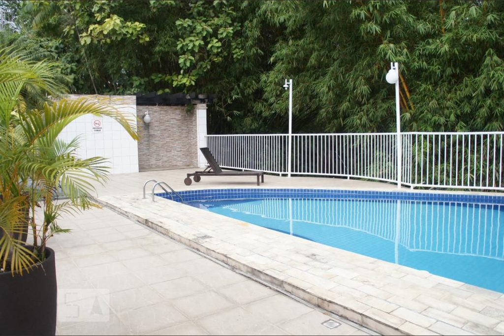 Piscina em um quintal com chão de pedras. Espreguiçadeira ao fundo