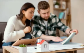 Imagem de um casal formado por um homem e uma mulher sentados no sofá da sala de um casa avaliando informações em um caderno que está em cima de uma mesa de centro ao lado de uma planta e uma casa de madeira em tamanho miniatura para ilustrar matéria sobre aluguel com título de capitalização