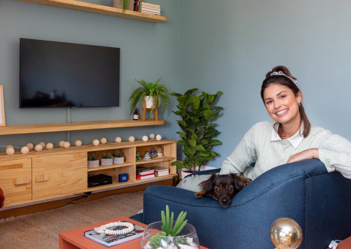 Foto que ilustra matéria sobre um apartamento no Jardim Prudência mostra a moradora do imóvel, Raissa Bóbbo, sentada em um sofá azul, com sua cachorrinha.