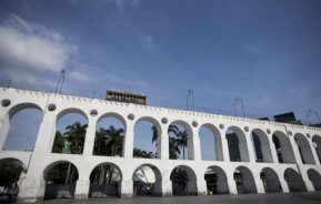 Foto que ilustra matéria sobre o Centro do Rio de Janeiro mostra a os Arcos da Lapa com o bondinho passando acima (Foto: Bruna Prado - MTUR)