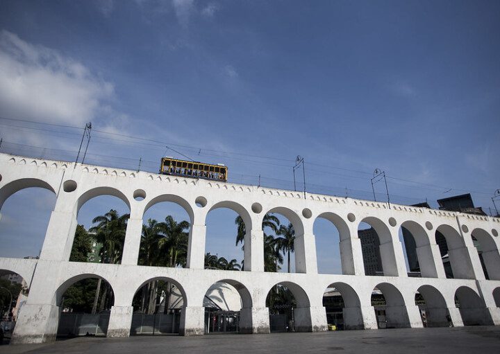 Foto que ilustra matéria sobre o Centro do Rio de Janeiro mostra a os Arcos da Lapa com o bondinho passando acima (Foto: Bruna Prado - MTUR)