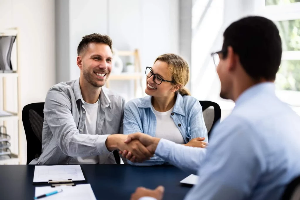 Imagem de um casal sorridente sentado em uma mesa em frente a um homem de terno, cumprimentando-o com um aperto de mão, em um ambiente de escritório moderno e bem iluminado