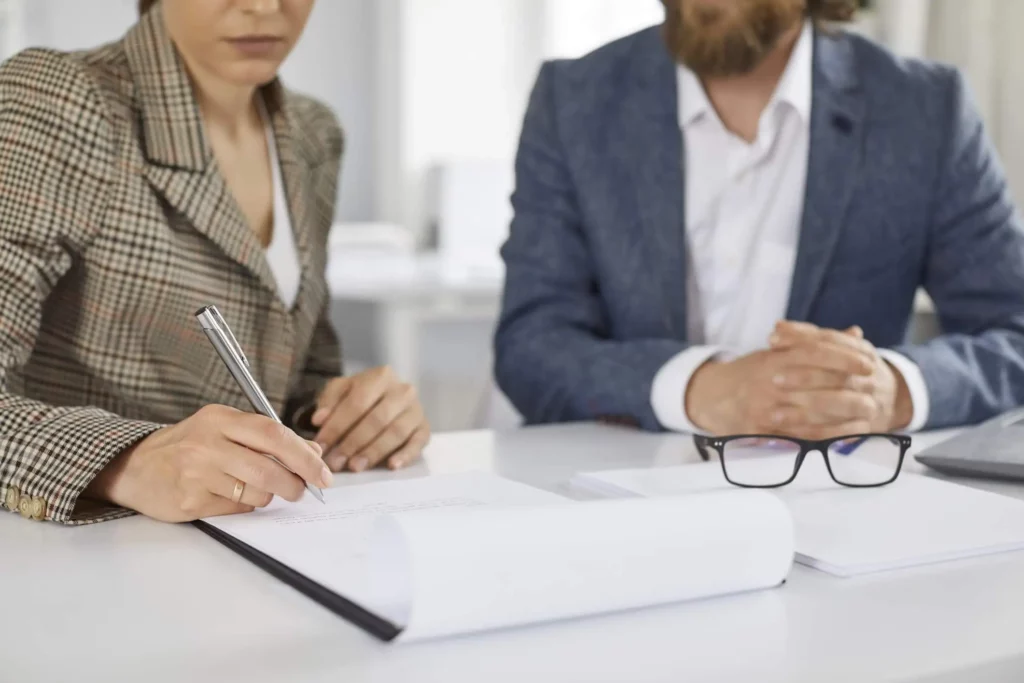 Imagem de uma mulher em um escritório assinando um documento ao lado de um homem de terno para ilustrar matéria sobre inventário de imóvel, quanto tempo demora?