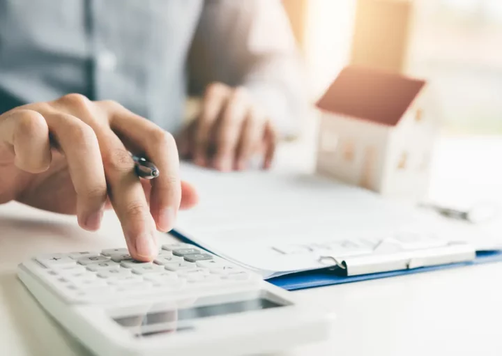 Homem analise documentos e realiza cálculos. Há uma miniatura de casa ao fundo da mesa.