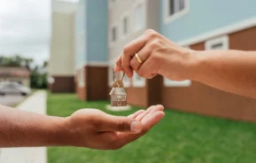 Imagem de uma pessoa com um anel de ouro entrega um chaveiro em forma de casa para outra em frente a um prédio de apartamentos e grama verde